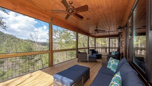 A covered outdoor deck with wooden flooring, wicker furniture, and a view of a forested landscape.