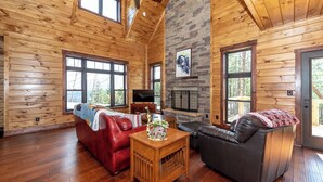 A log cabin interior with a fireplace, leather sofas, a TV, and large windows.