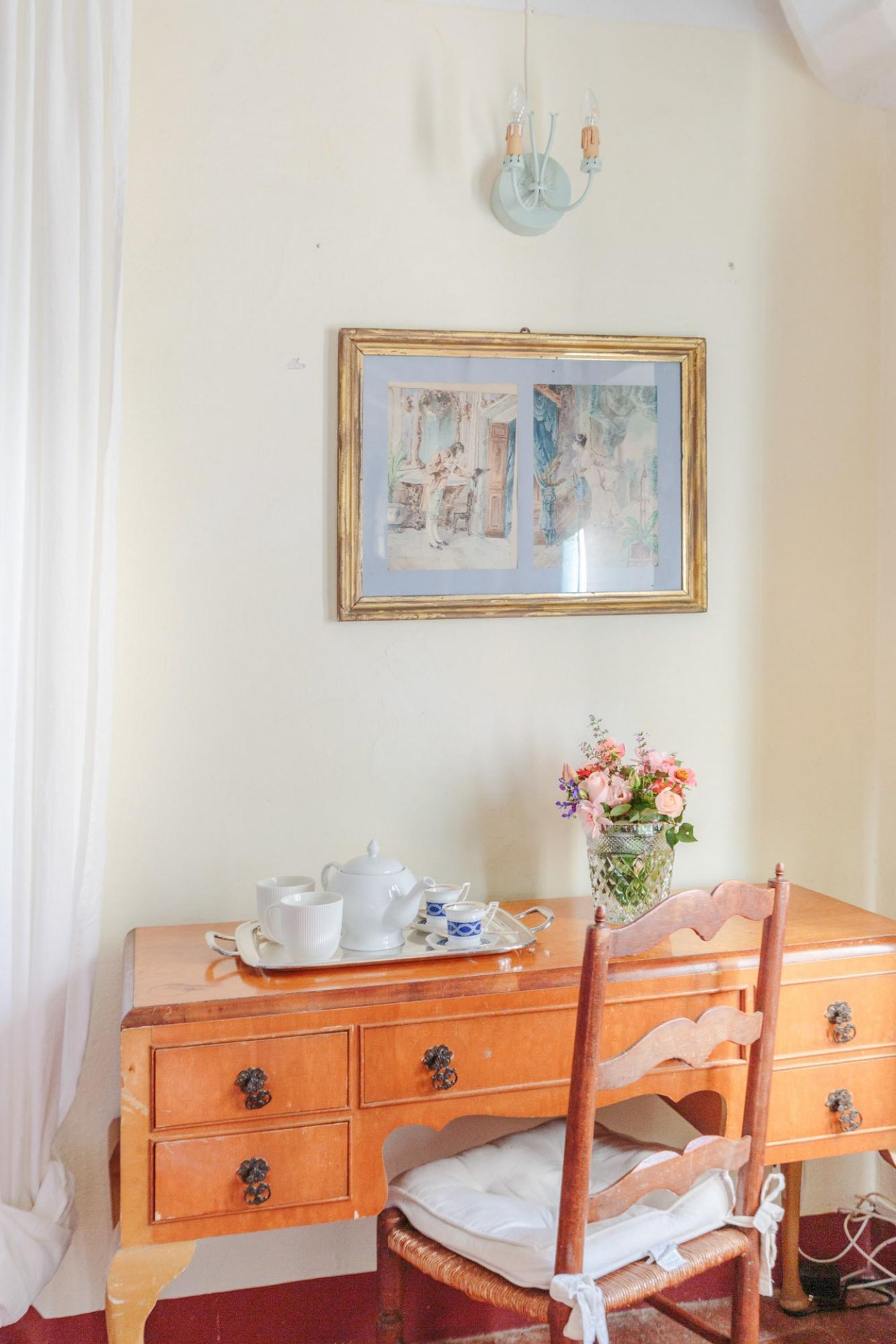 A wooden desk with a white chair, a framed picture on the wall, a vase with flowers, and a tea set on the desk.