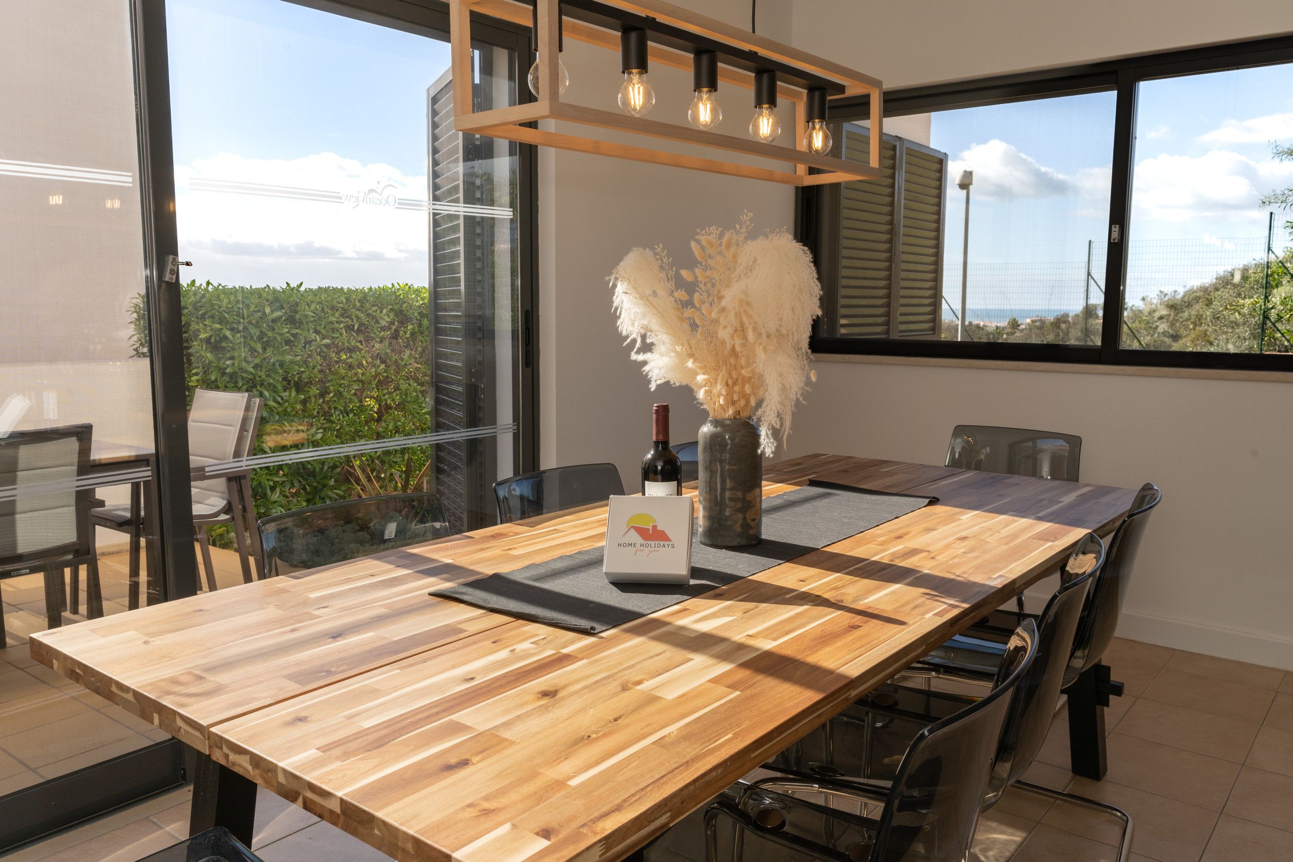 Un comedor moderno con una mesa de madera, sillas, un jarrón con plumas de pampa y una botella de vino.
