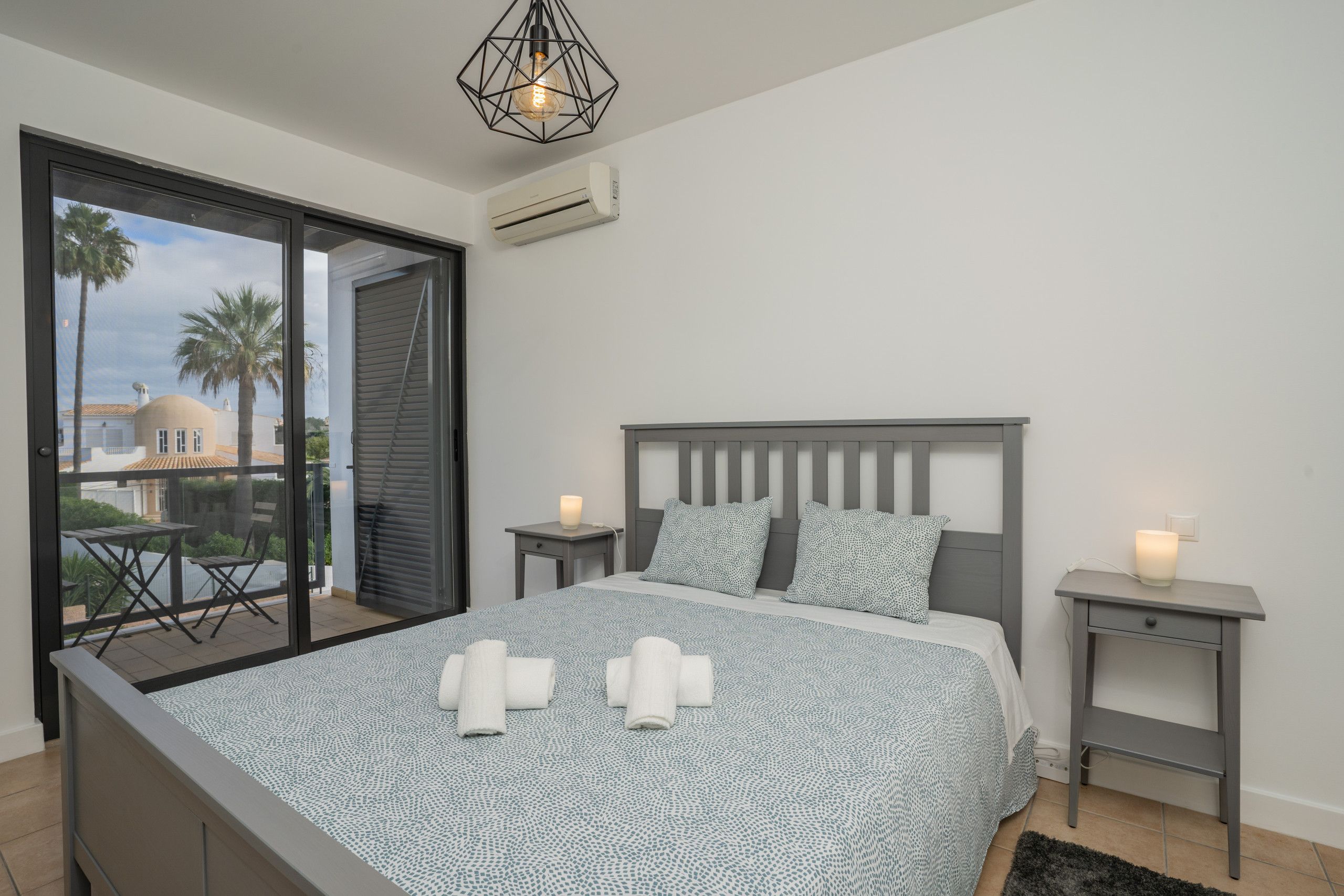 A bedroom with a bed, two bedside tables, a nightstand, a ceiling light fixture, and a view through a sliding door.