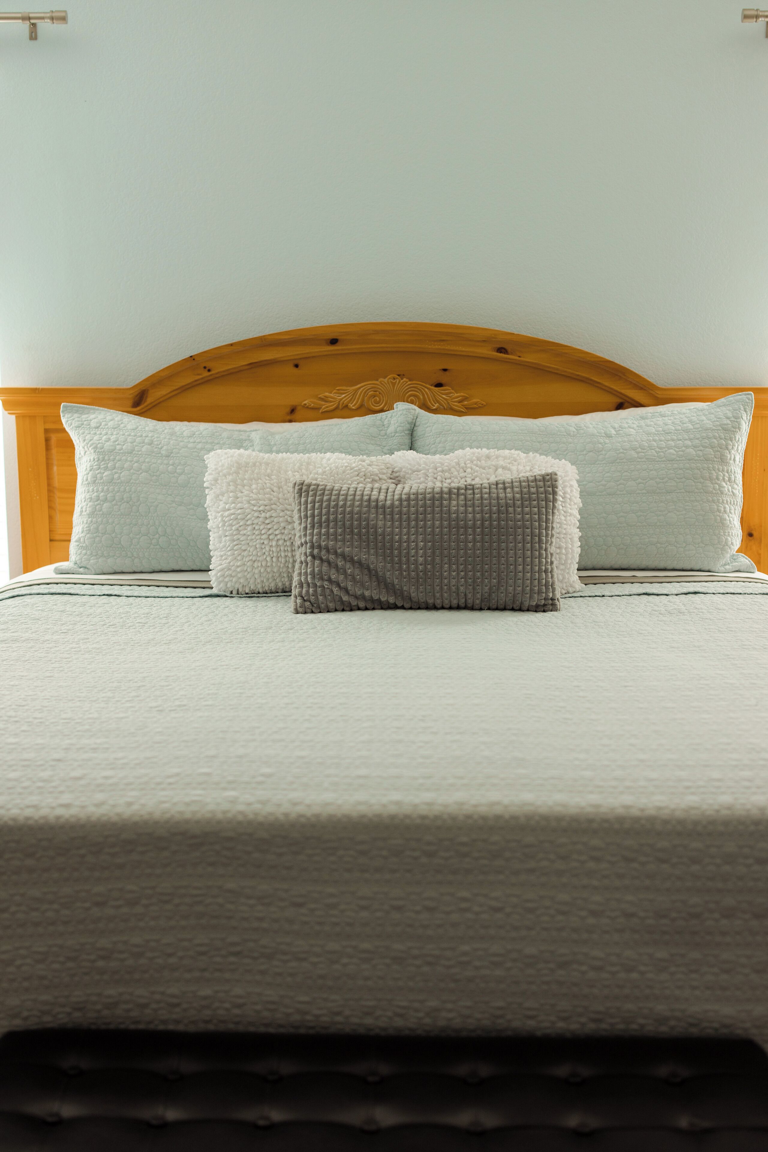 A neatly made bed with a wooden headboard, a light-colored bedspread, and three pillows.