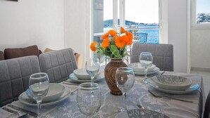 A dining table set with plates, glasses, and cutlery, a vase with flowers, and a view of the sea through large windows. - Apartments Happy Holiday (Rogoznica)