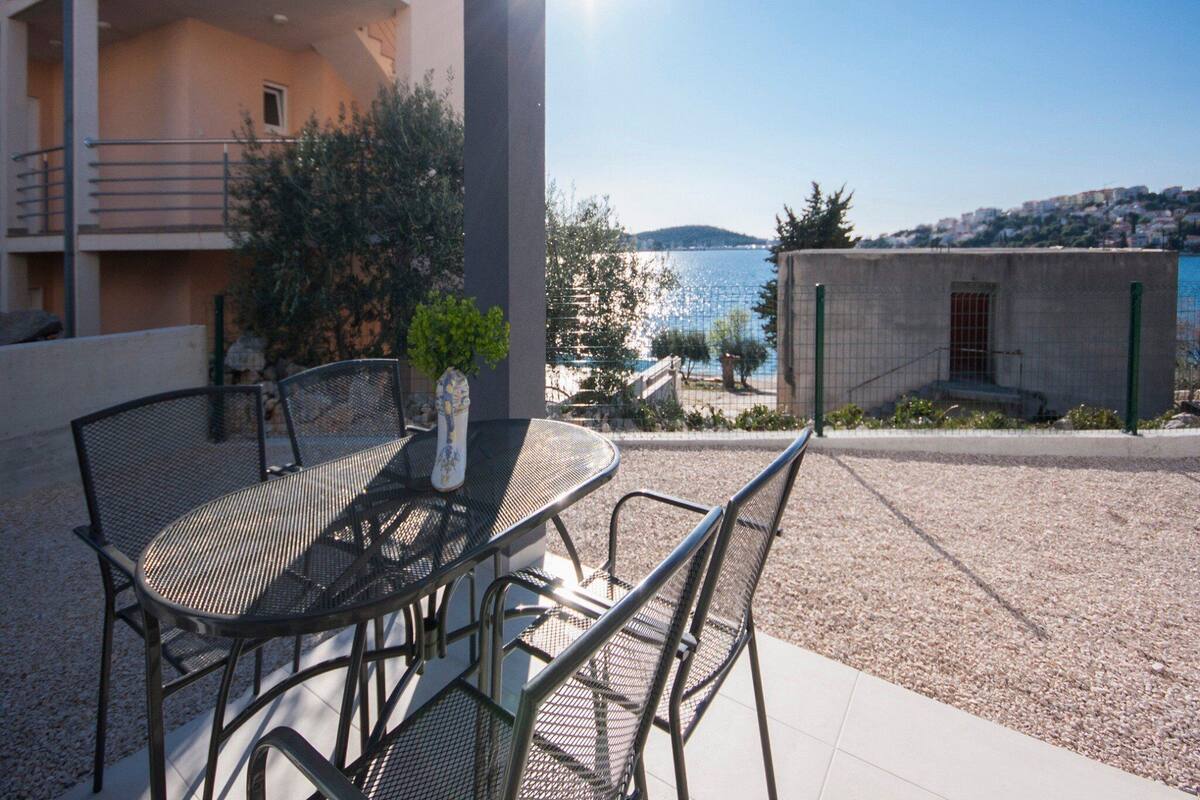 Un espace extérieur aménagé avec une table et des chaises, un pot de fleurs, et une vue sur la mer et les bâtiments en arrière-plan.