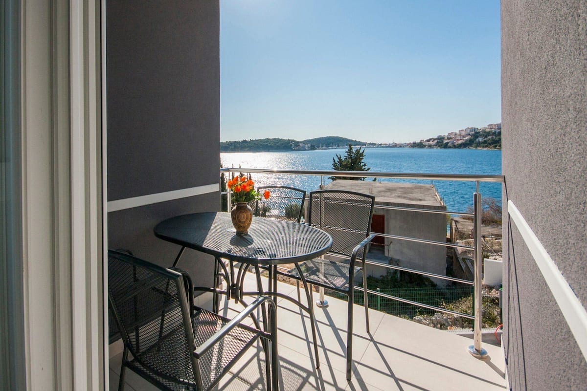 Un balcon avec une table et des chaises, un vase rempli de fleurs et une vue sur une étendue d’eau et des collines lointaines.