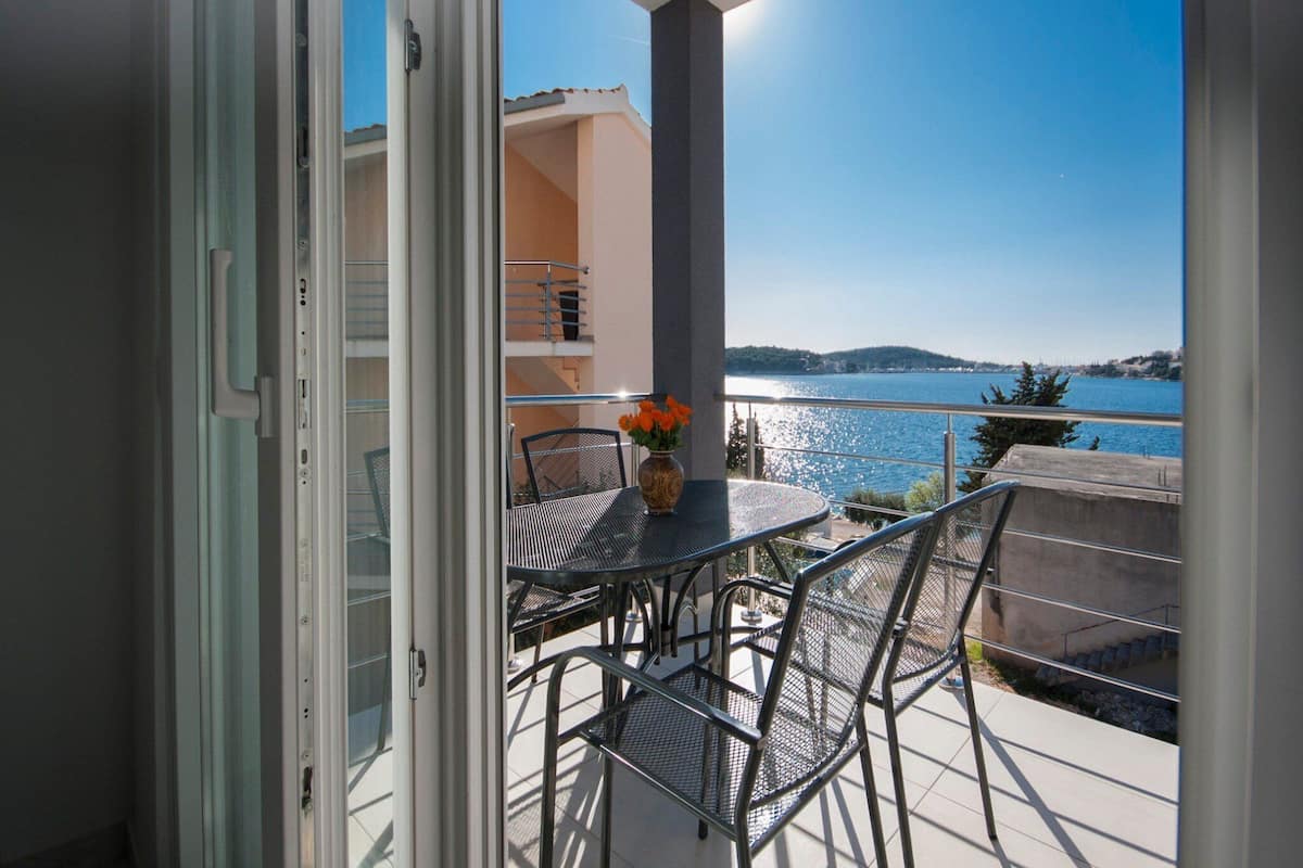 Un balcon ouvert avec une table et des chaises, un vase rempli de fleurs et une vue sur une étendue d’eau et des collines lointaines.