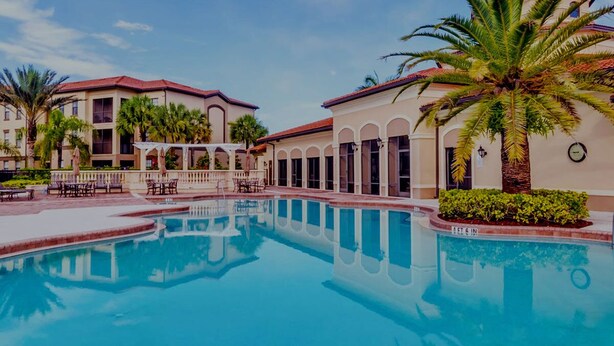 A swimming pool with a clear blue water surface, surrounded by palm trees and a building with a red-tiled roof. - Beautiful Naples Retreat! (Naples)