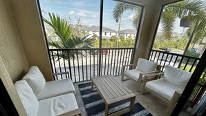 A balcony with white cushioned seating and a wooden table, overlooking a residential area with houses and palm trees. - Beautiful Naples Retreat! (Naples)