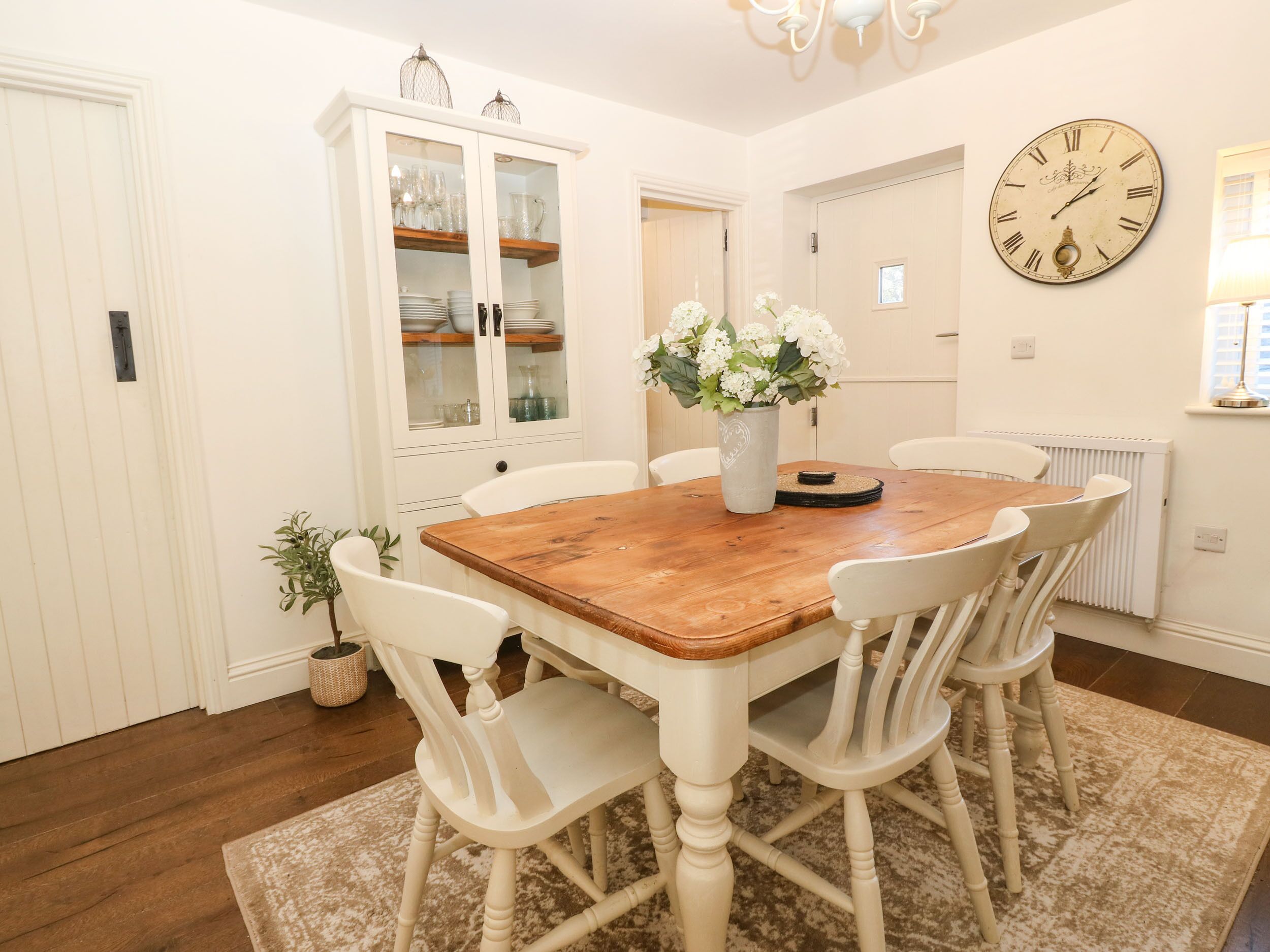 A dining room with a wooden table, white chairs, a white cabinet with glass doors, and a wall clock.