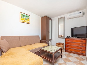 Una habitación con un sofá marrón, una mesa de centro de vidrio, un aparador de madera con cajones, un televisor, un espejo y un equipo de aire acondicionado.