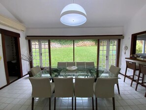 Uma área de jantar com mesa de vidro e seis cadeiras, grandes janelas com vista para um gramado e um lustre.