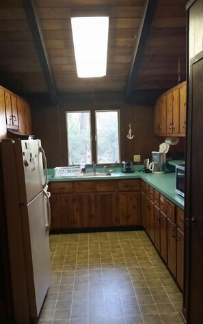 Una cocina con armarios de madera, un refrigerador, un lavamanos y un ventanal con vista a los árboles.