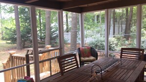 Une terrasse couverte avec une table et des chaises en bois, un panier en osier rempli de serviettes et une vue sur une zone boisée.