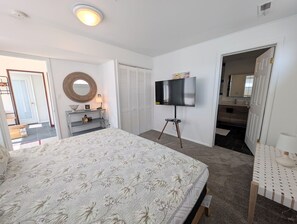 Ein Schlafzimmer mit einem großen Bett, einem Fernseher auf einem Schrank, einem Spiegel und einer Tür zu einem weiteren Raum.