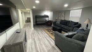 Un salon moderne avec un canapé gris, un téléviseur fixé au mur et un lit superposé à l’arrière-plan.