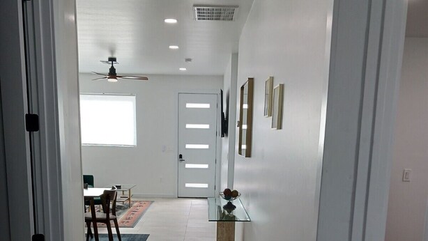 A modern hallway with a dining area in the background, featuring a ceiling fan, recessed lighting, and a door with a window.