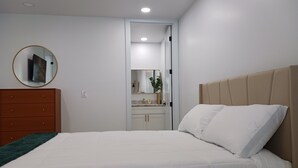 A bedroom with a bed, a dresser, a mirror, and a bathroom visible through an open door.