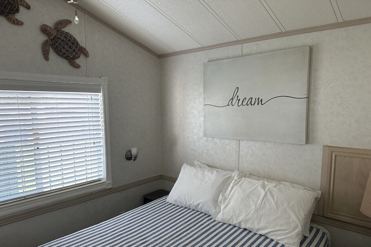 Ein Schlafzimmer mit einem Bett, einem Fenster mit Jalousien, einer Wandleuchte und einem gerahmten Wort 'dream' an der Wand.