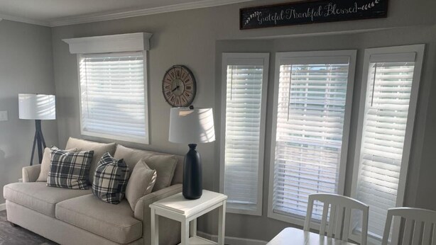 A living room with a beige sofa, white table, and chairs. There are three windows with blinds, a clock, and a sign that says "Grateful Thankful Blessed." - Caribbean Shores Vacation Rentals (Jensen Beach)