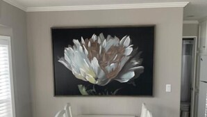 A dining area with a white table and chairs, a large floral painting on the wall, and a kitchen in the background. - Caribbean Shores Vacation Rentals (Jensen Beach)