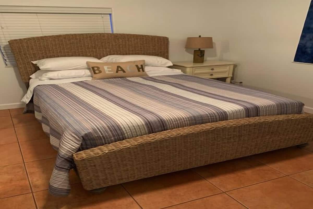 Ein Bett mit gestreiftem Bettbezug, ein Kissen mit dem Schriftzug "BEACH", ein Nachttisch mit Lampe und ein Fenster mit Jalousien.