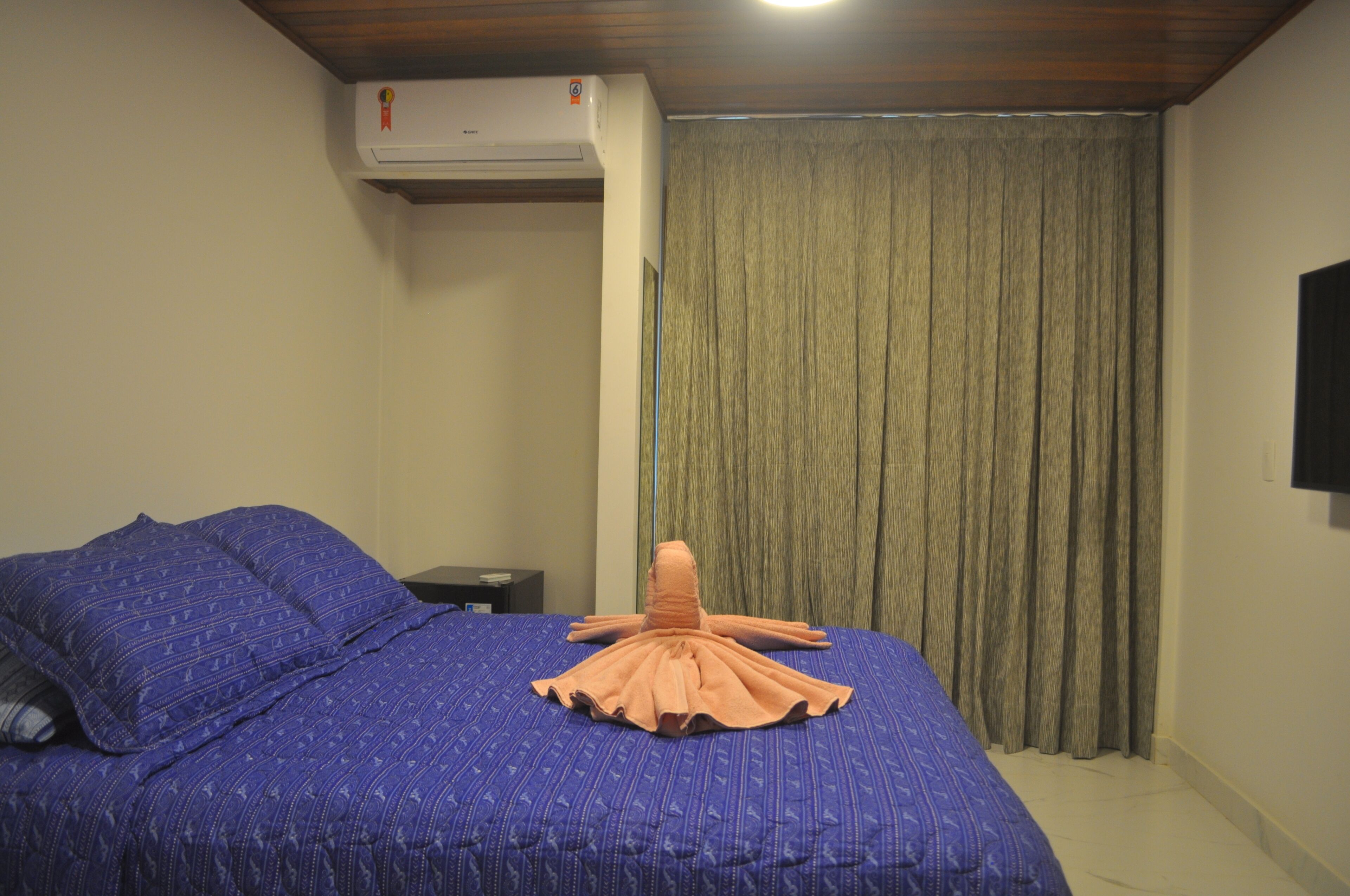 A hotel room with a bed, a nightstand, a wall-mounted air conditioner, and a window with curtains.
