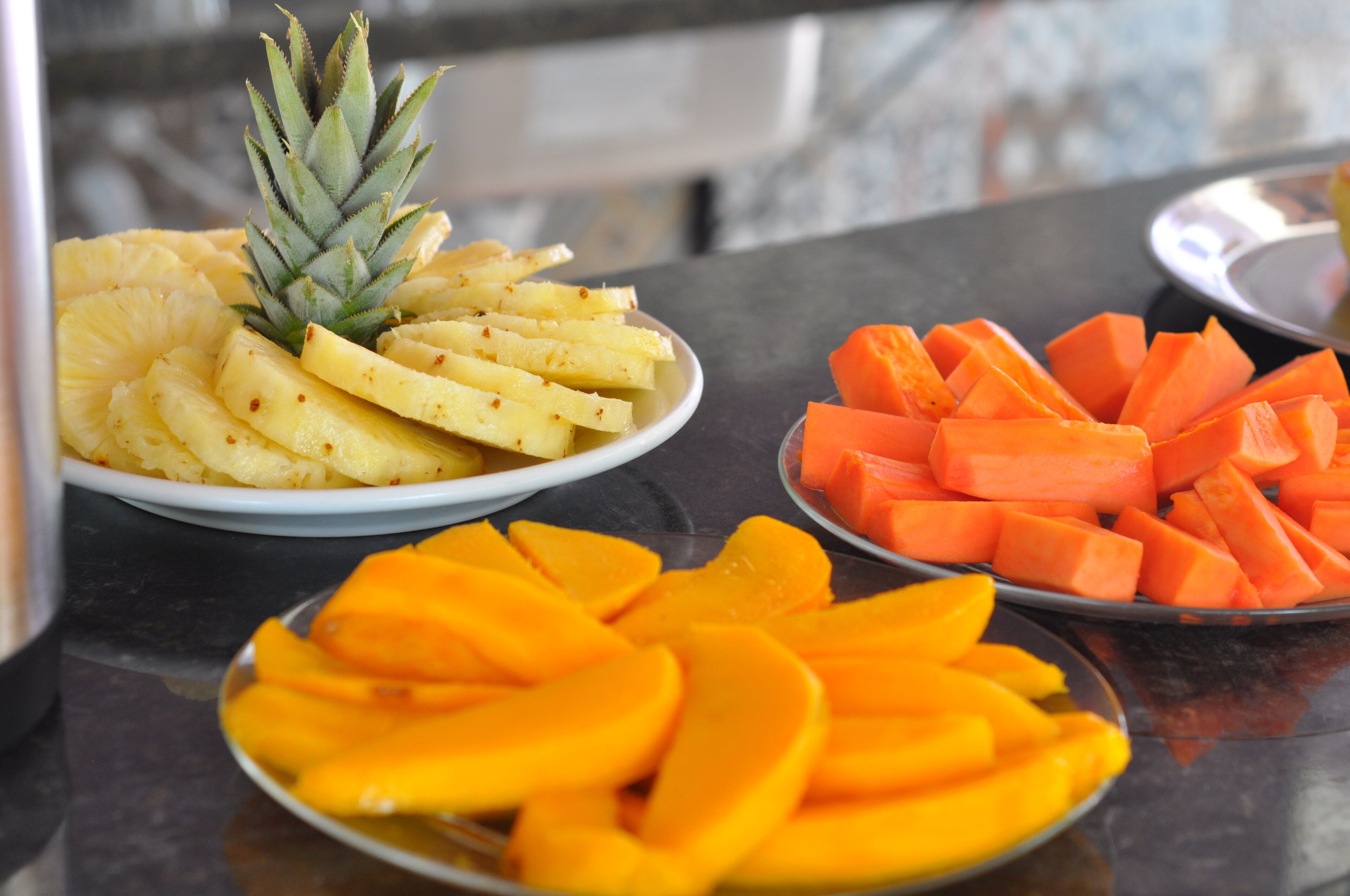 Una mesa con tres platos de frutas cortadas: piña, mango y zanahoria.