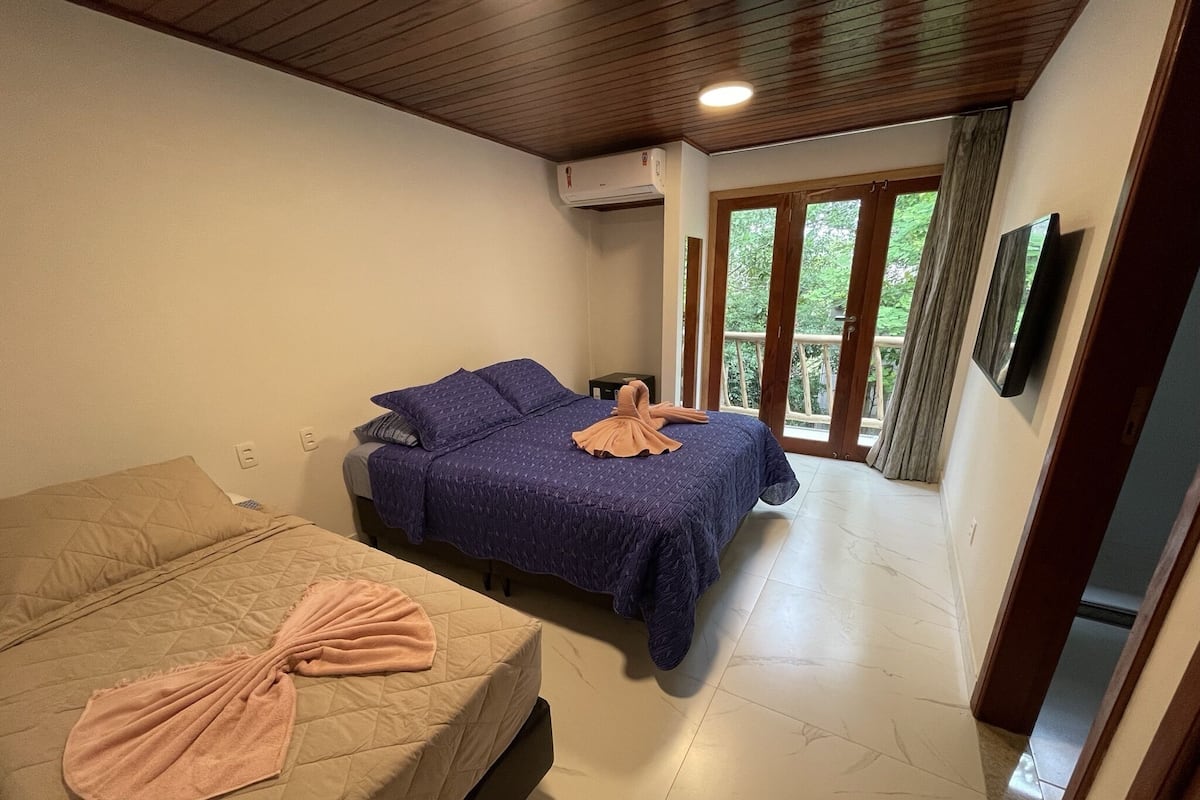 A bedroom with two beds, a TV, and a balcony with a view of greenery.