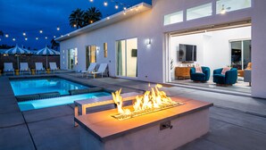 A modern outdoor pool area with a fire feature, lounge chairs, and a well-lit interior space.