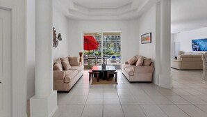 A spacious living room with two beige sofas, a coffee table, and a large window with a red umbrella. - Tipsy Turtle - Beautifully Landscaped Secluded pool and spa home (Venice)