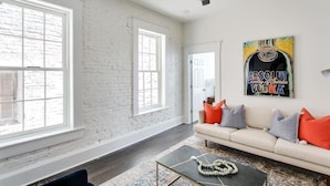 A spacious living room with a white brick wall, large windows, a ceiling fan, and a sofa with colorful cushions.