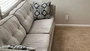 Un sofá color beige con almohadones estampados, piso alfombrado y una ventana con persianas.