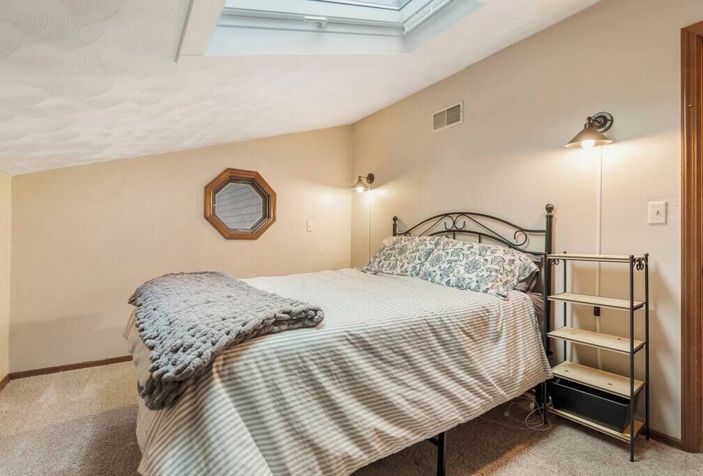 Un dormitorio con cama, tragaluz, estante montado en la pared y lámpara de noche.