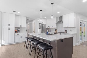 A modern kitchen with white cabinetry, a large island with bar stools, stainless steel appliances, and pendant lighting. - Airy, Spacious, Newly Renovated Home – Walk to UF! (Gainesville)