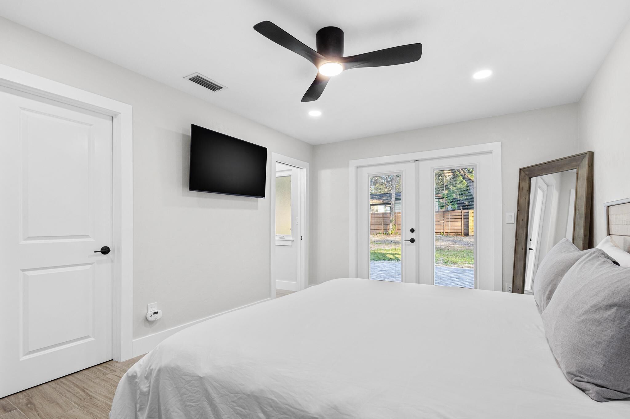 Un dormitorio con cama, ventilador de techo, televisor montado y puerta corrediza de vidrio que da a un patio trasero.