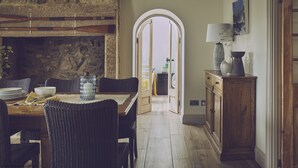 Un comedor con chimenea de piedra, una mesa de madera con sillas, vista a través de un arco y un aparador con lámpara.