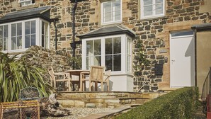 A stone house with a bay window, a front door, and a small porch with a table and chairs. - Driftwood -  a pet friendly that sleeps 4 guests  in 2 bedrooms (Craster, Alnwick)
