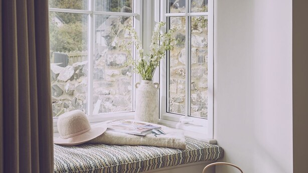 A room with a large window, a wicker basket, a white vase with flowers, and a hat on a bench. - Driftwood -  a pet friendly that sleeps 4 guests  in 2 bedrooms (Craster, Alnwick)