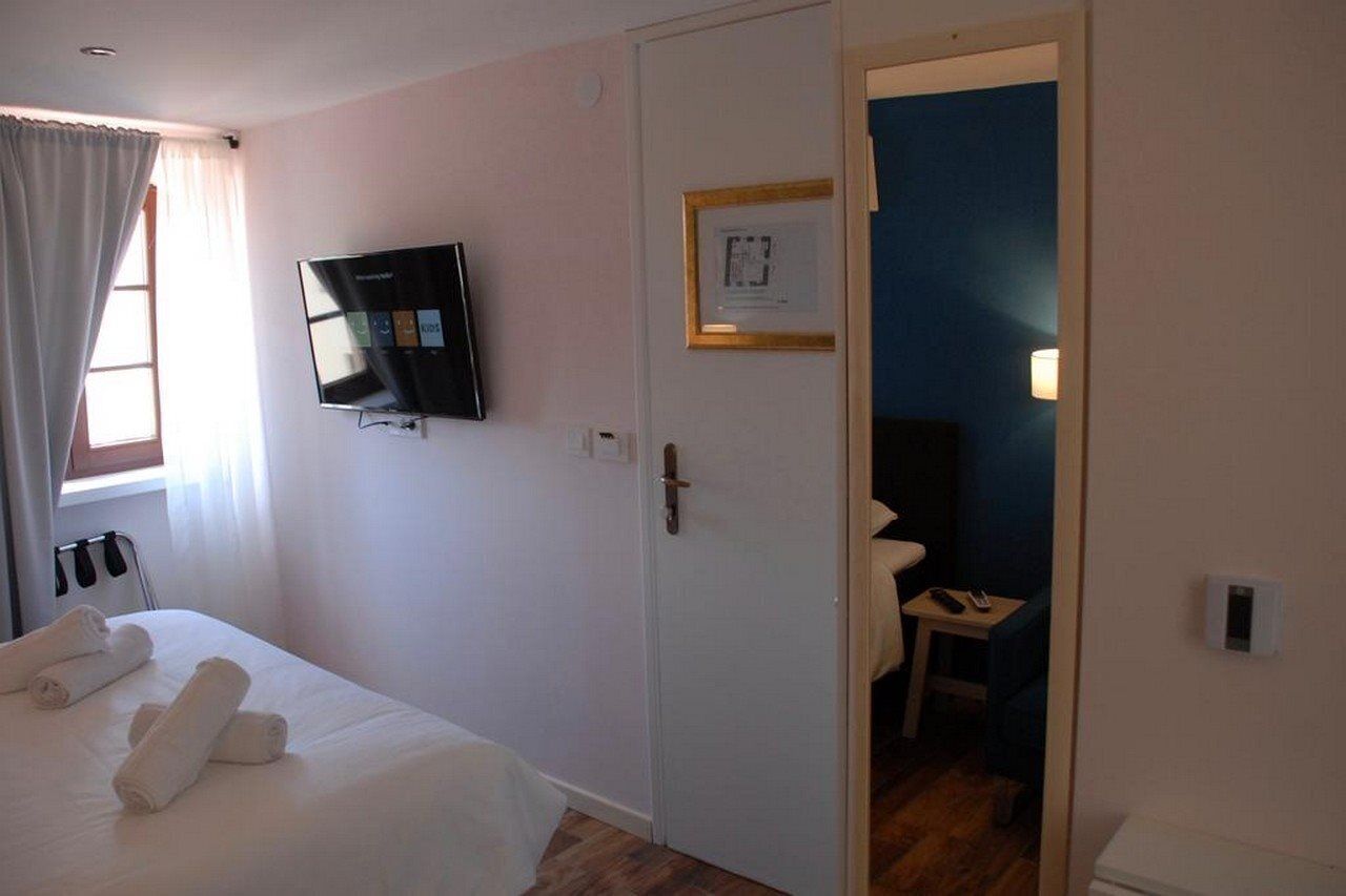 A hotel room with a bed, a TV mounted on the wall, a door leading to another room, and a small table with a lamp.