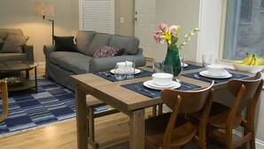 Ein Essbereich mit einem Holztisch, gedeckt für vier Personen, umgeben von Stühlen. Im Hintergrund befindet sich eine Couch und eine Stehlampe.