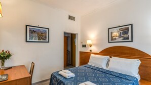 A hotel room with a bed, a desk, a chair, a lamp, and two framed pictures on the walls. - Baltic Hotel (Rome)