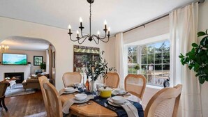 A dining area with a table set for four, surrounded by wooden chairs. A chandelier hangs above the table, and a large window provides a view of the outdoors. - Charming cottage in the city; walk to Main Street (Vancouver)