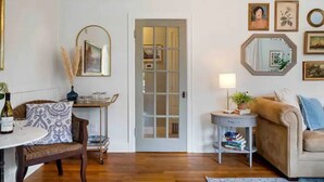 A living room with a wooden floor, a beige sofa, a small round table with a lamp, a chair, and a bookshelf. - Charming cottage in the city; walk to Main Street (Vancouver)