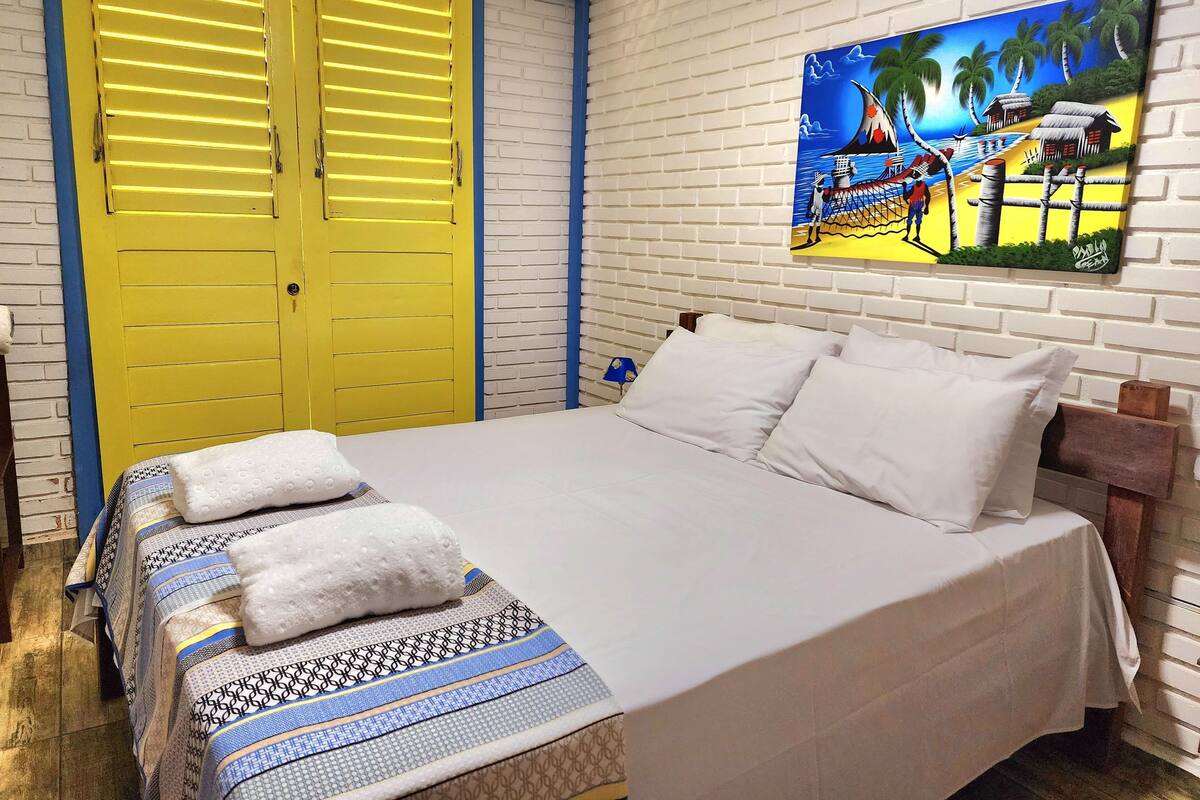 A hotel room with a bed, a painting of a beach scene, and a yellow door.