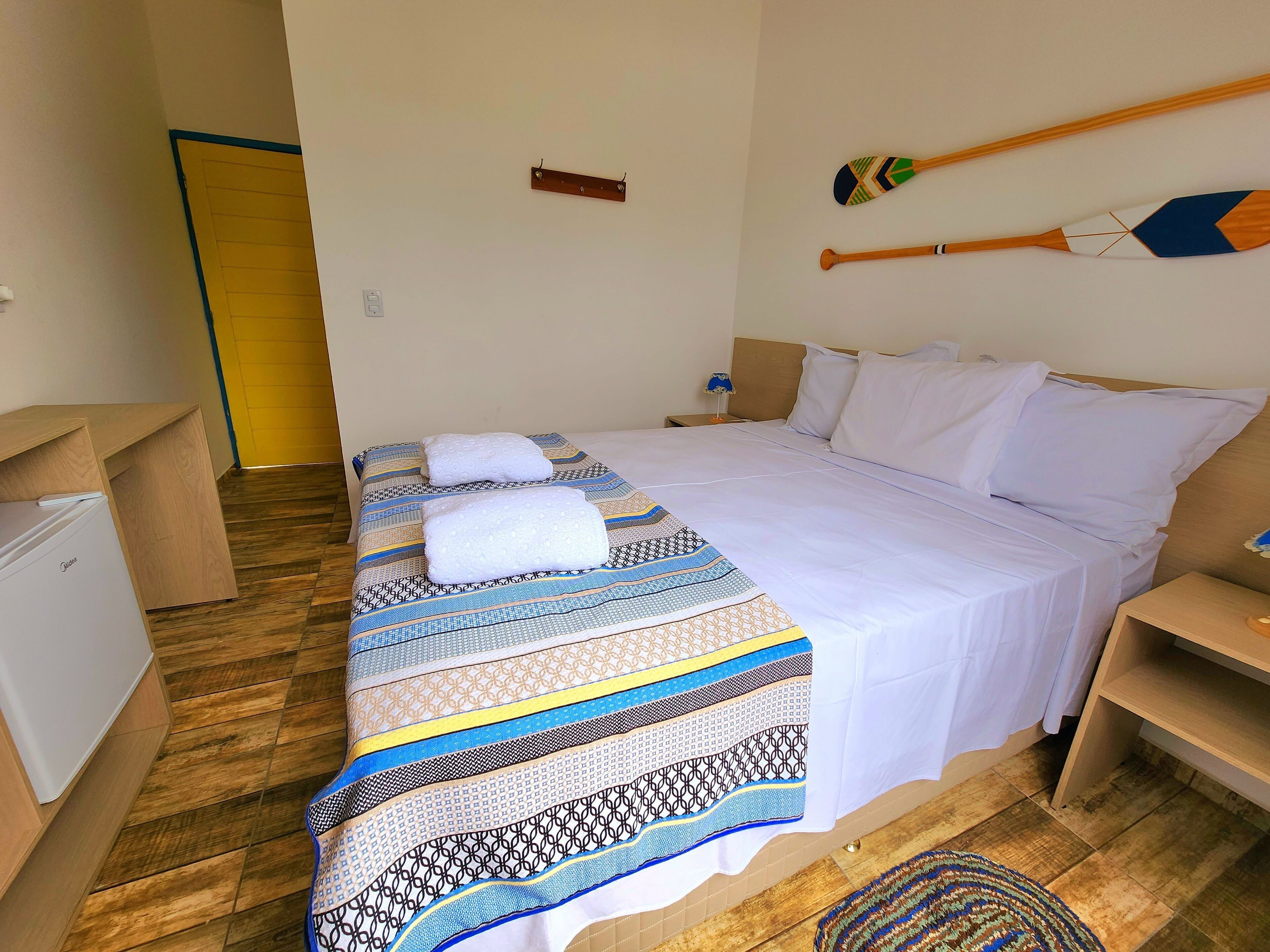 A neatly made bed with patterned bedding, a bedside table, a small wooden nightstand, and a wall-mounted wooden paddle.