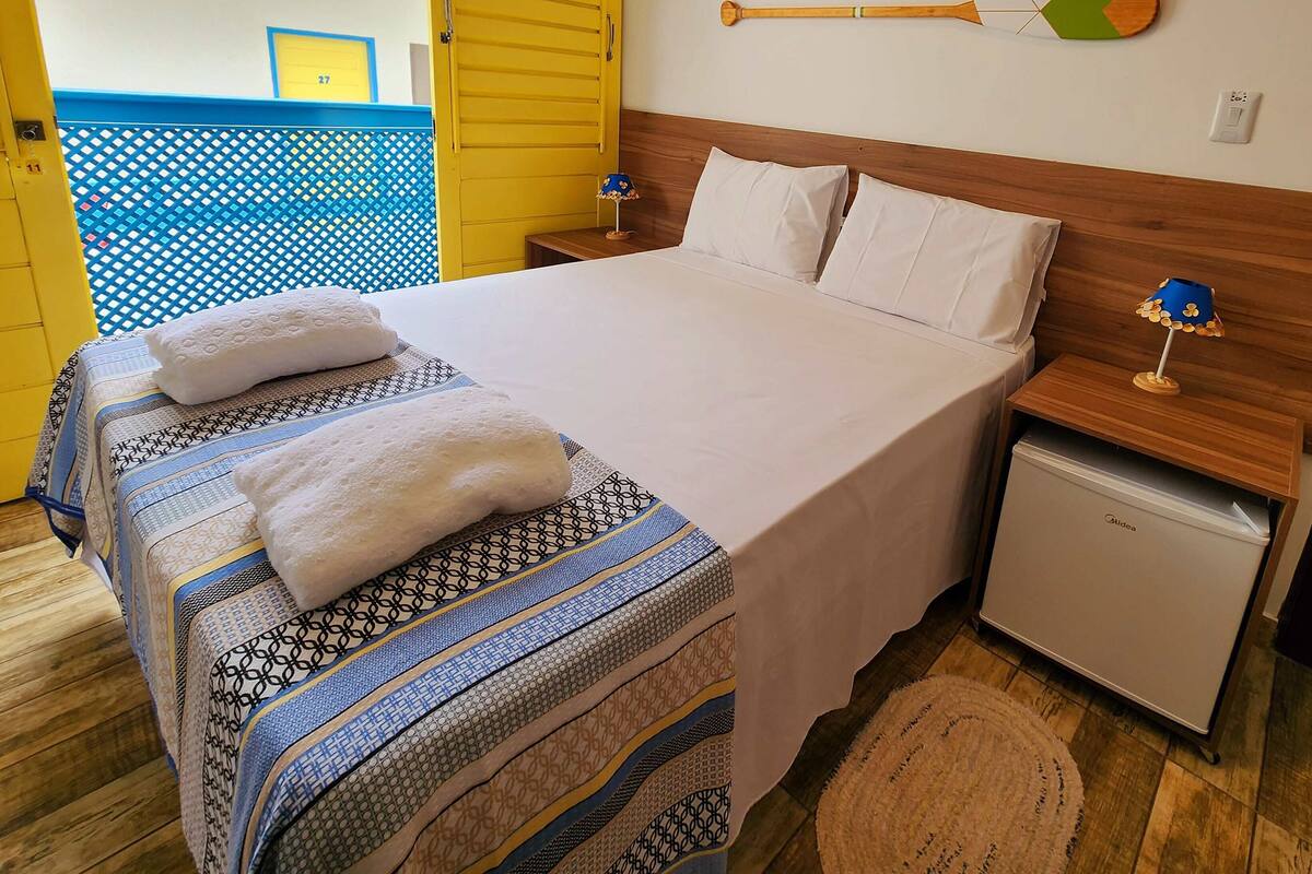 A bedroom with a bed, bedside tables, a nightstand, a small refrigerator, and a wall-mounted paddleboard.