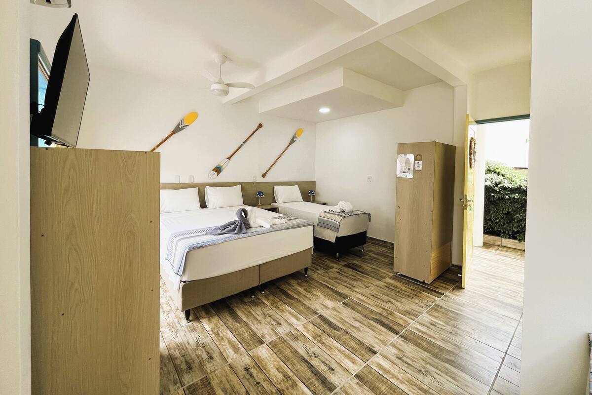 A hotel room with a wooden floor, two beds, a ceiling fan, and a view of greenery outside.