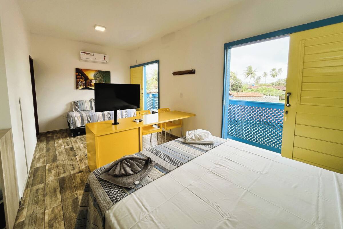 A hotel room with a bed, a desk with a chair, a television, and a window with a view of palm trees.