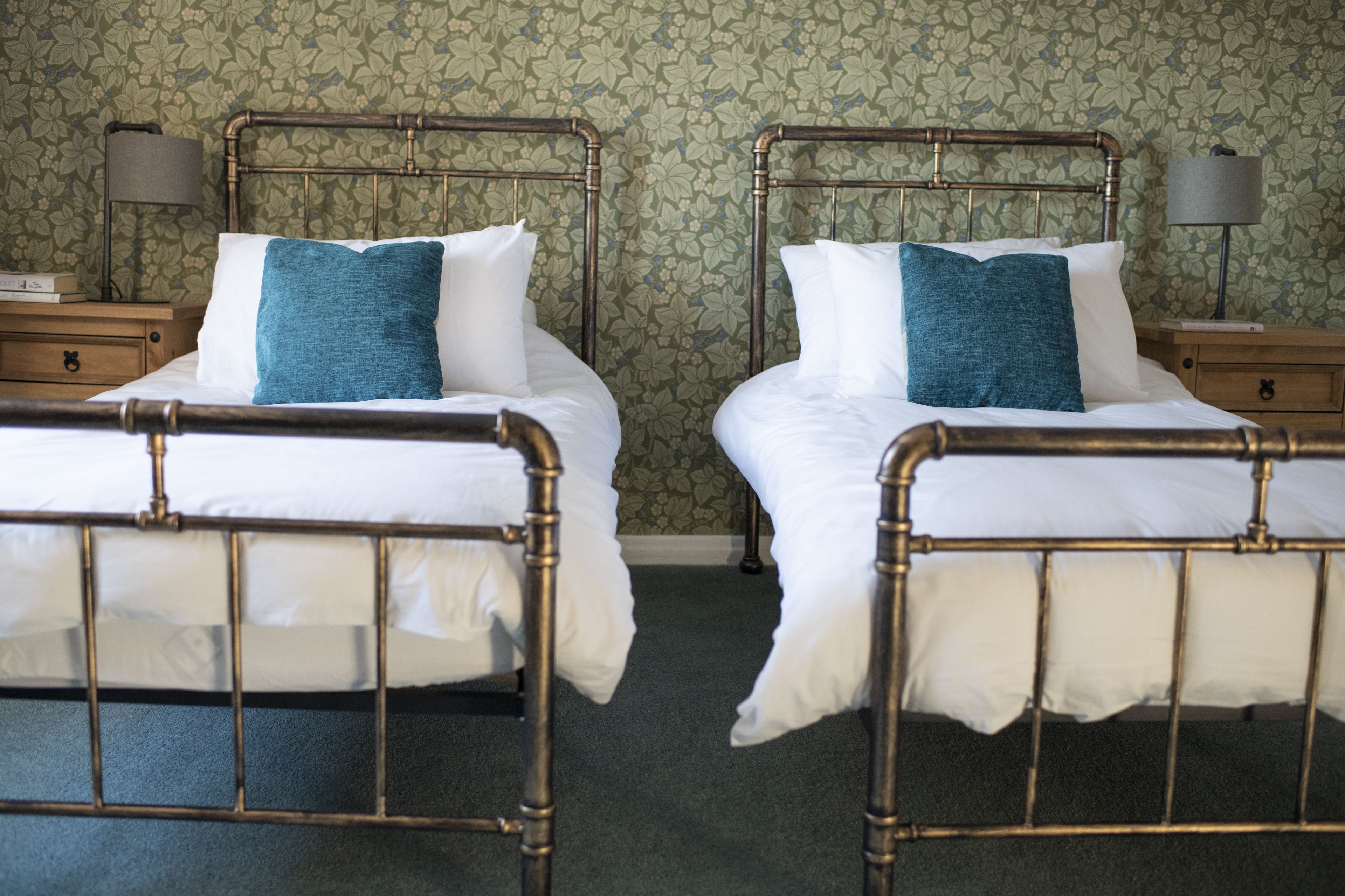 Two twin beds with metal frames, white bedding, and teal pillows. Each bed has a wooden nightstand with a lamp. The room has floral wallpaper.