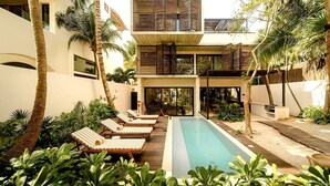 A modern outdoor pool area with lounge chairs, palm trees, and a two-story building with large windows. - Azumaya Beach House Tulum – Beachfront, Chef Included (Tulum)